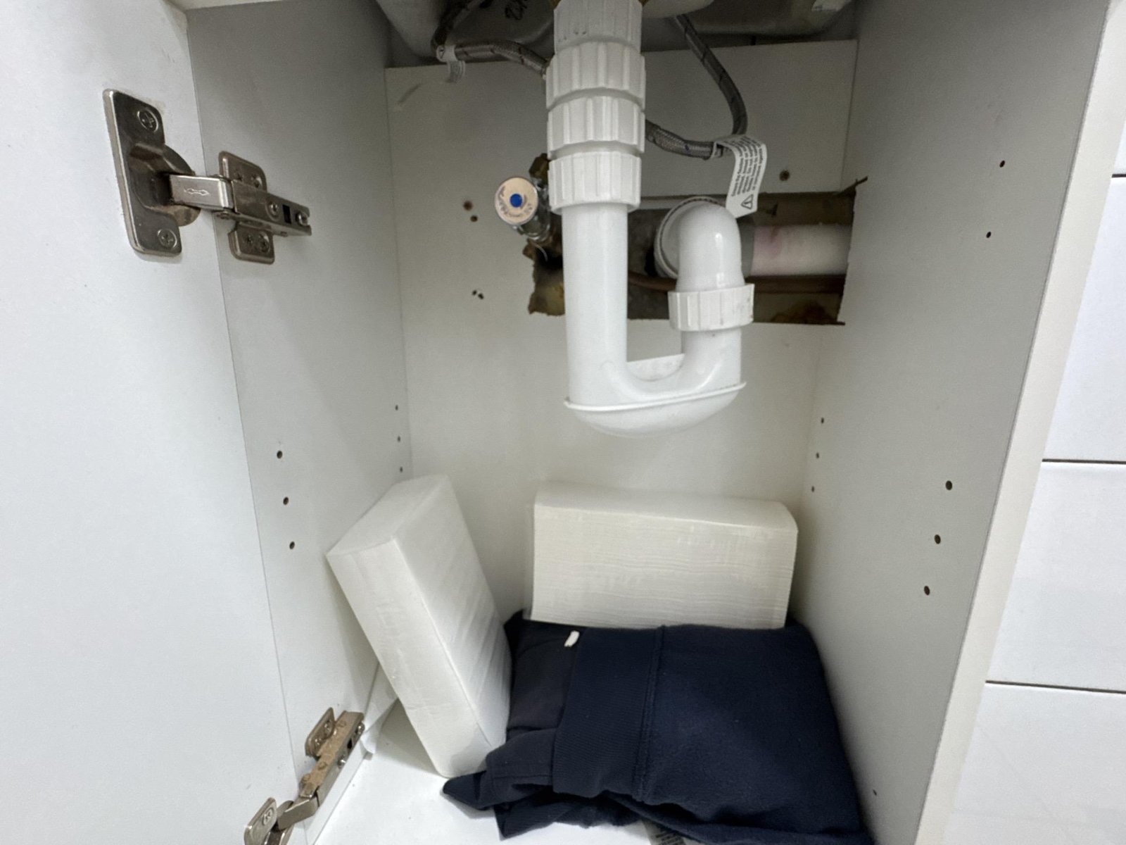 Inside a vanity cupboard, a white sink trap and pipework with bagged towels and cloth on the floor.