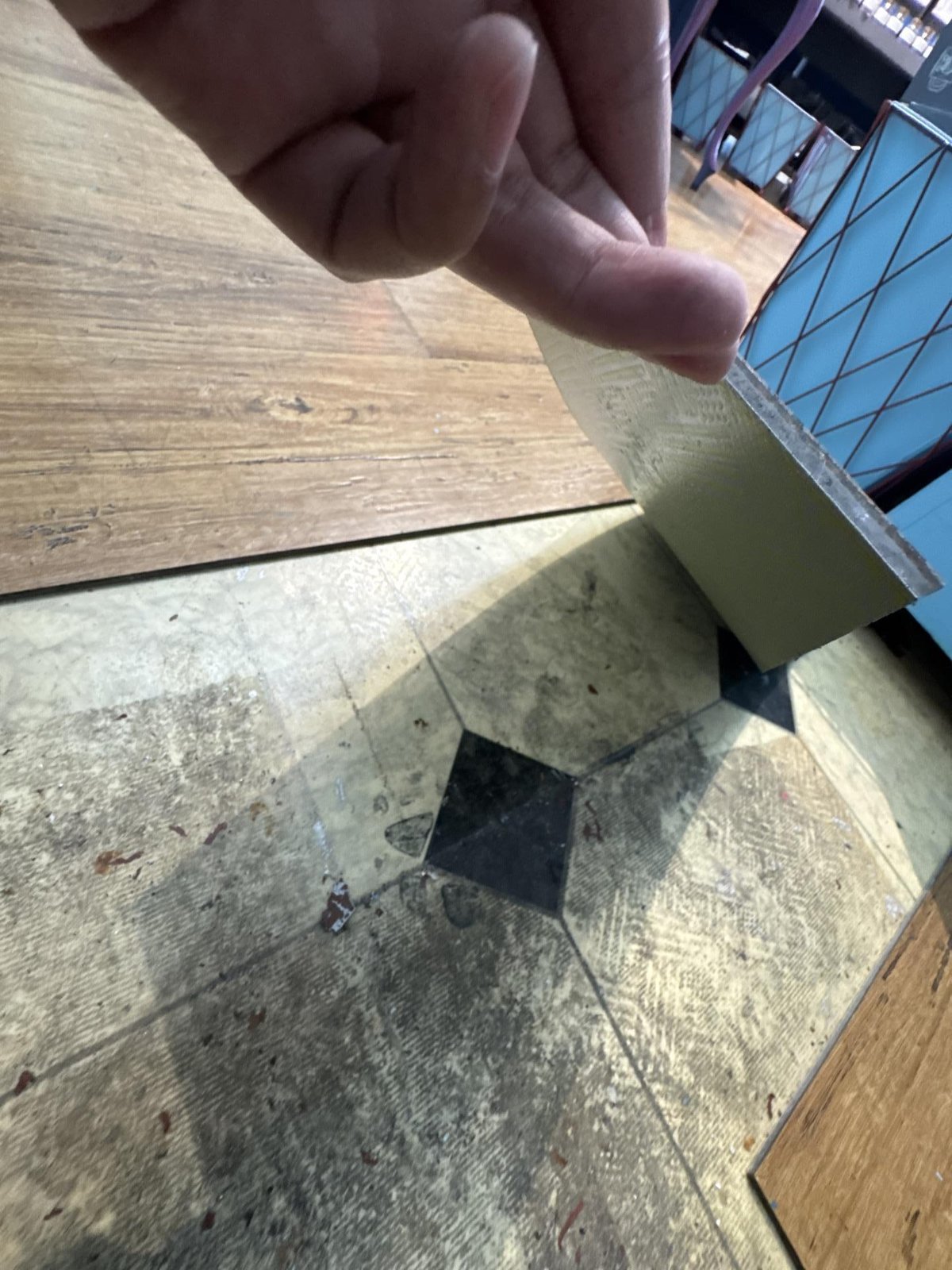 A hand holds a sample of a sheet flooring tile against a worn, stained floor.