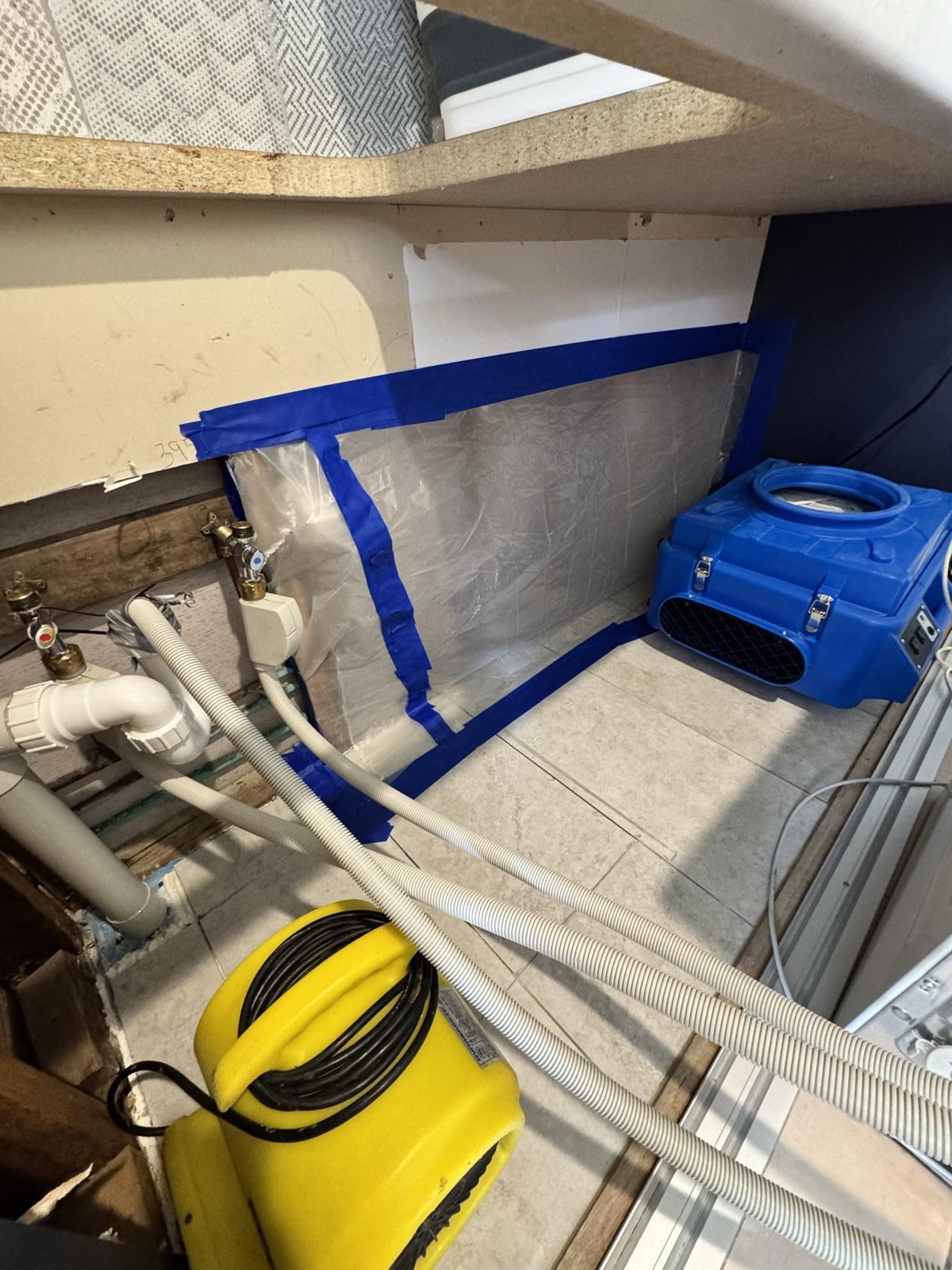 Blue air mover and yellow equipment with hoses drying a tiled floor behind taped plastic sheeting.