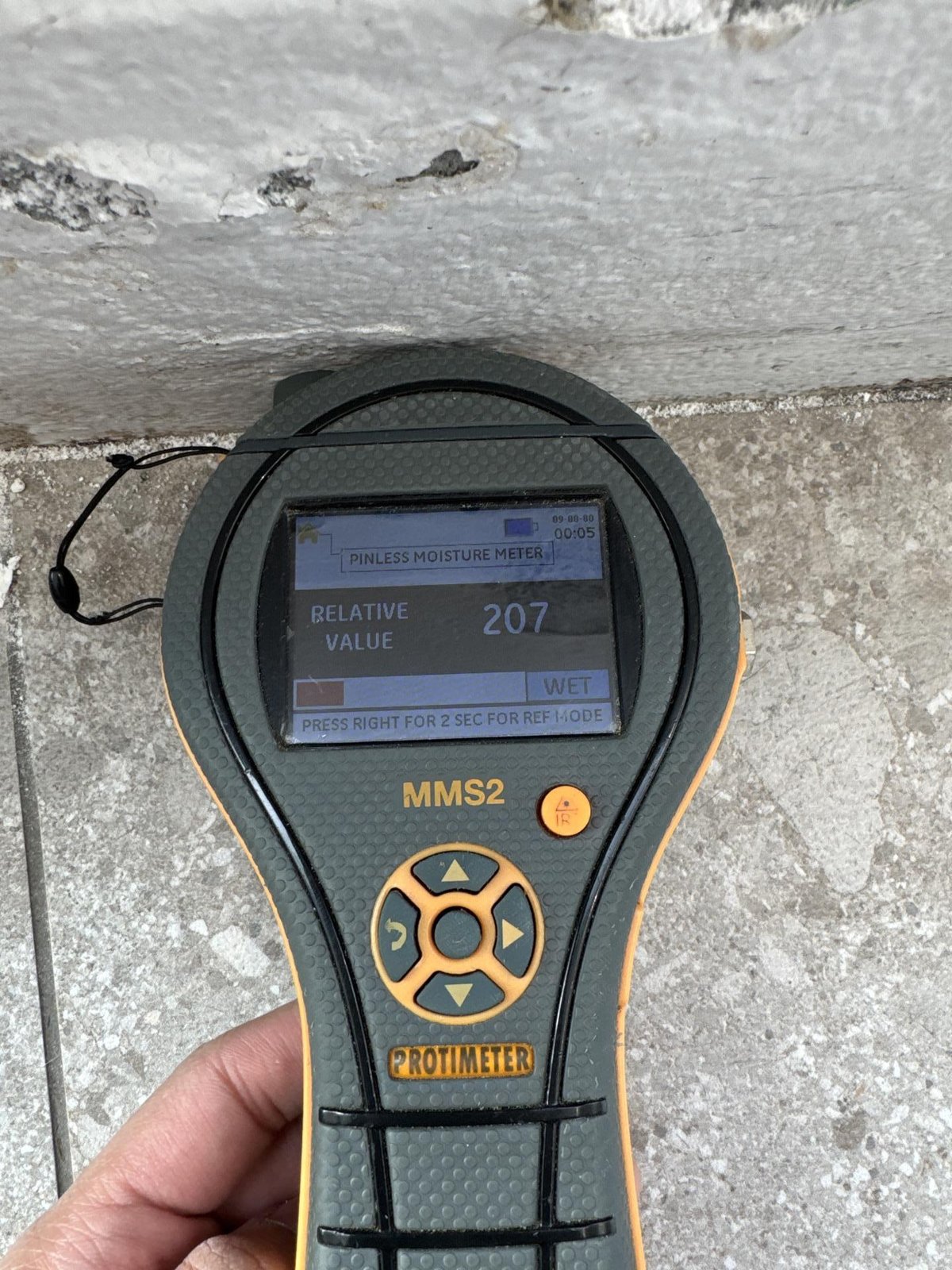 A hand holding a Protimeter moisture meter against a wet, stained concrete wall