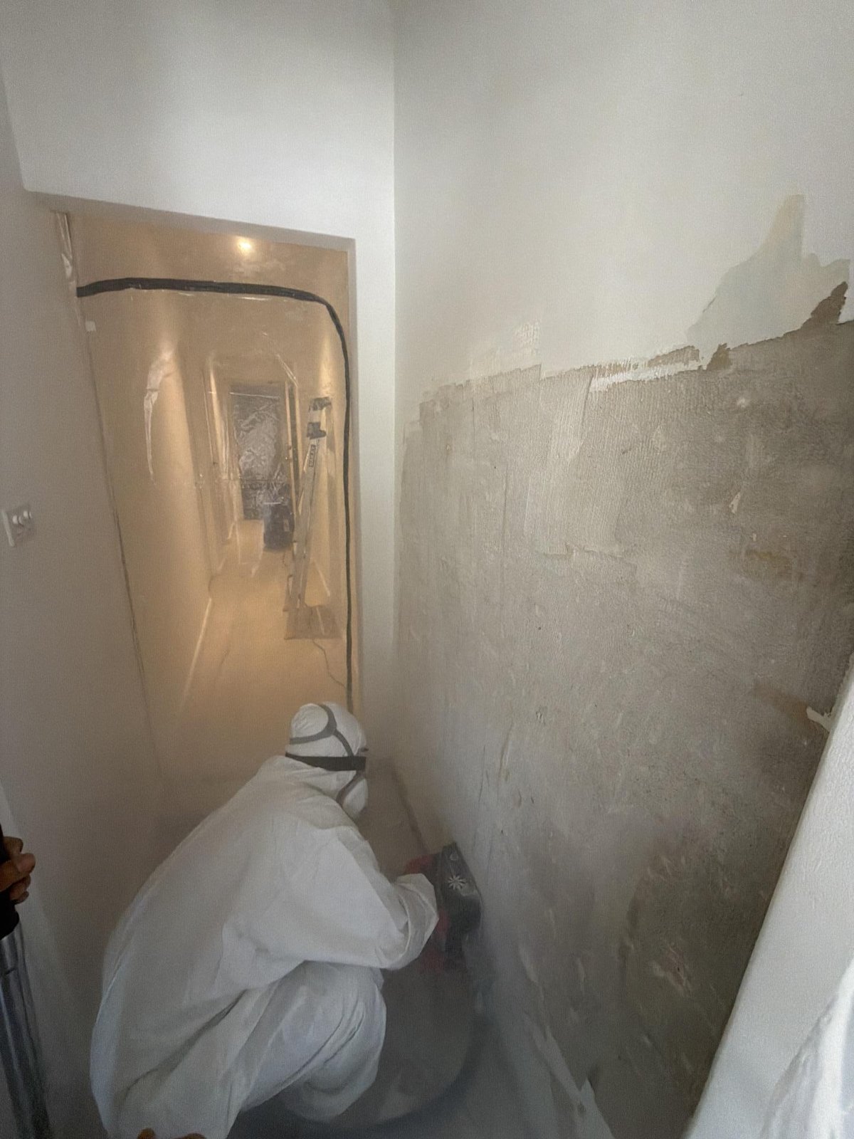 A technician in white protective suit and respirator sanding a wall in a plastic-sheeted doorway, with stripped plaster showing.