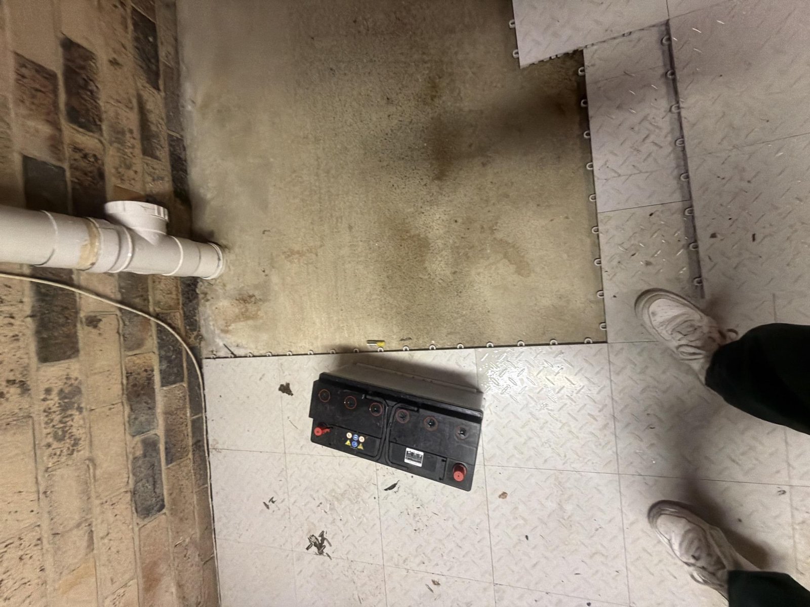 A stained concrete floor with a black battery and PVC pipe, beside a diamond-plate floor.