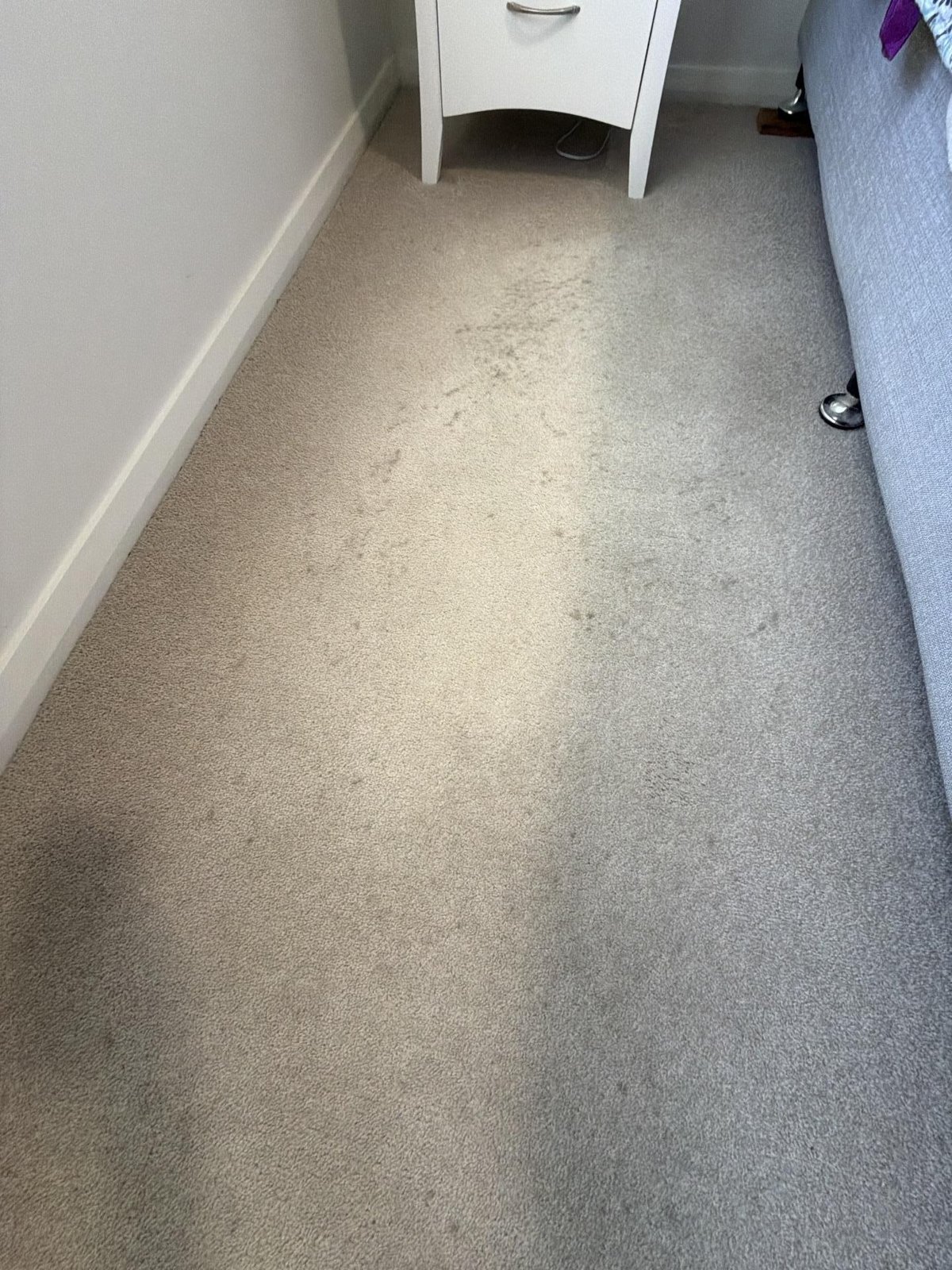 Stained carpet beside a bedside cabinet, with faint darker marks across the pile.