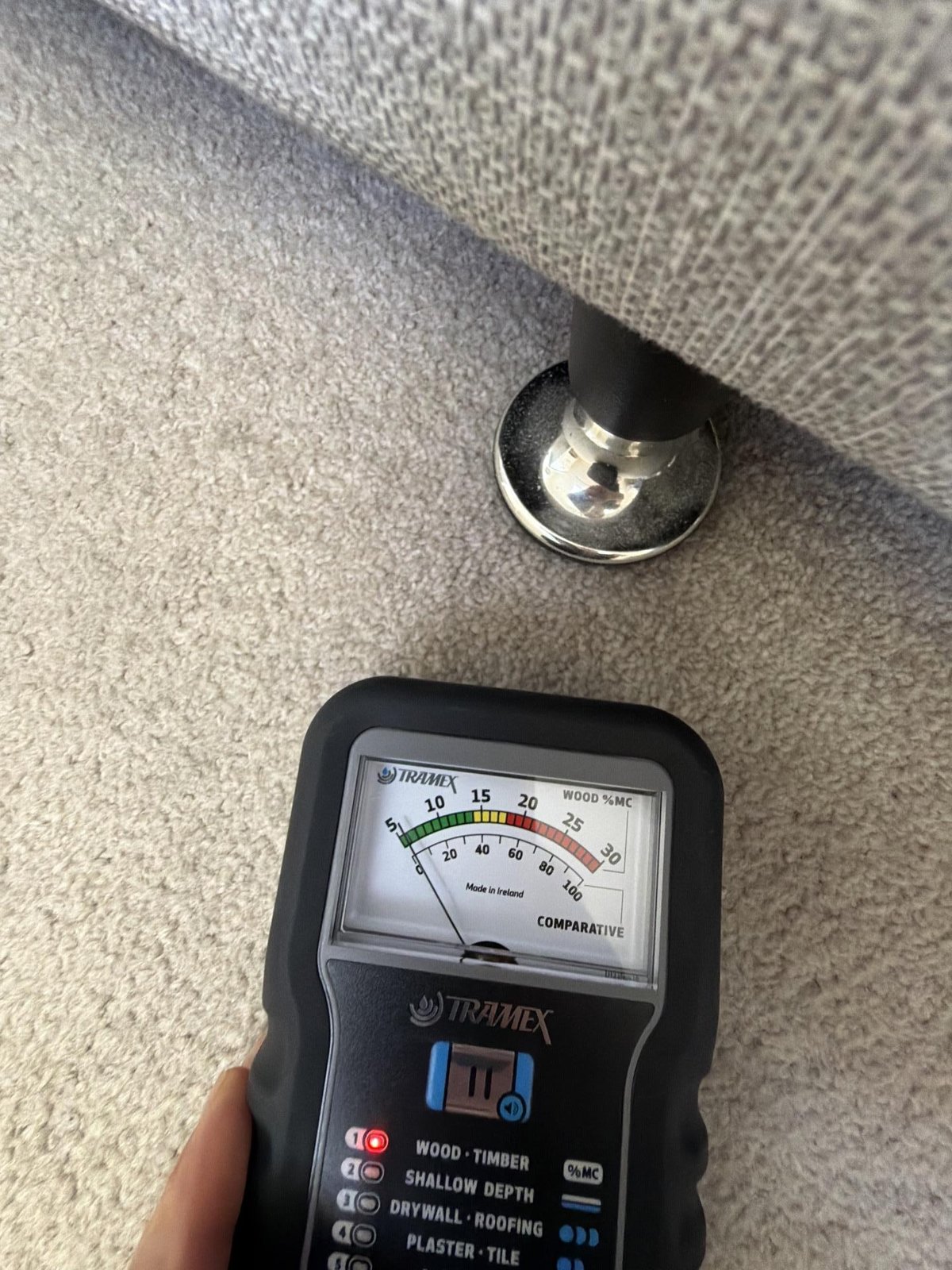 A technician's moisture meter held against carpet beside a chrome-based skirting or stair fixture