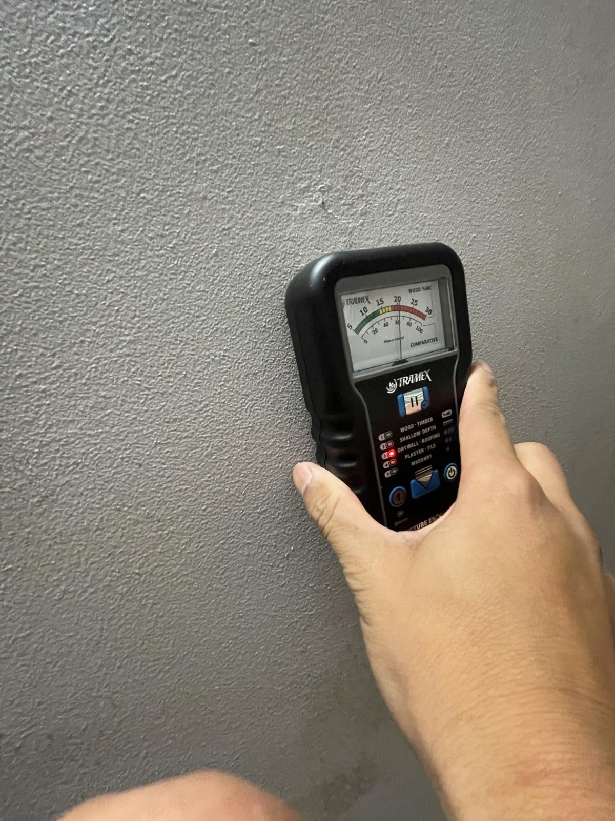 A hand holds a moisture meter against a grey wall to test for dampness.