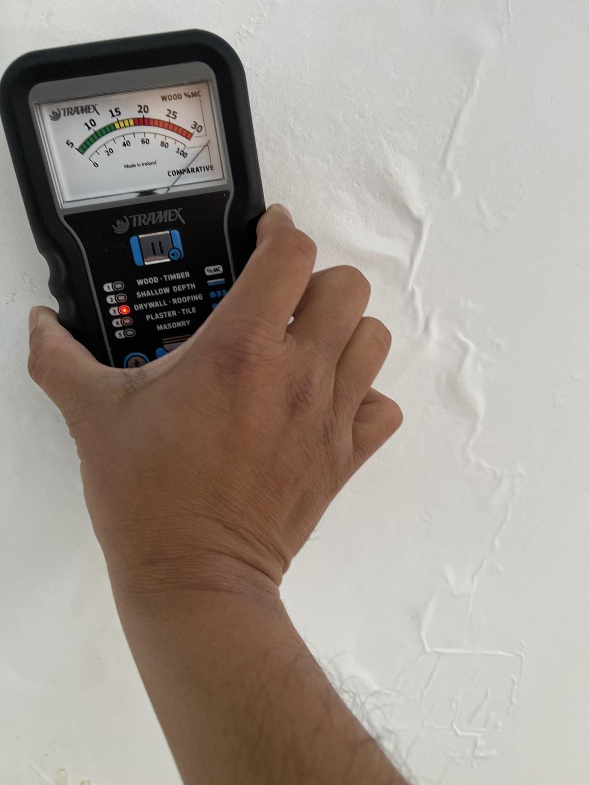 A technician's hand holding a moisture meter against a textured white wall.