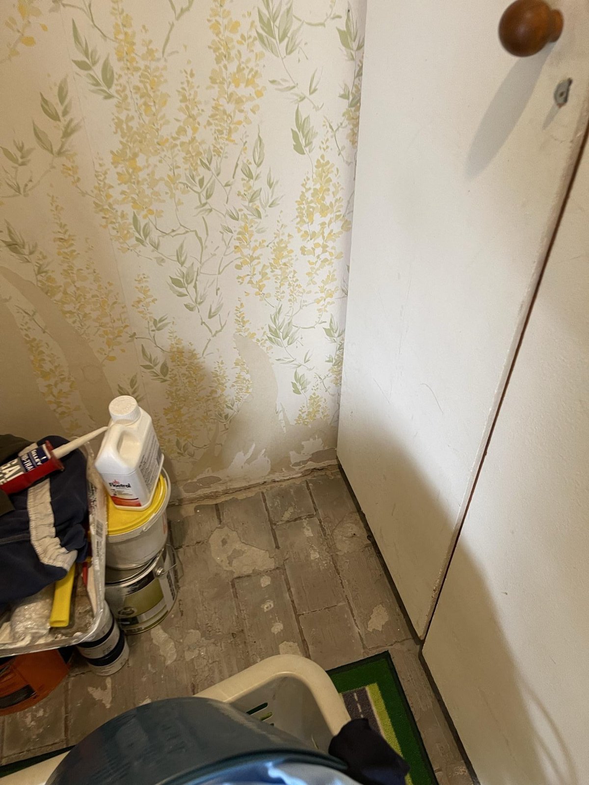 A small room with stripped floorboards and lifted wallpaper, with paint tins and a bucket beside the wall.
