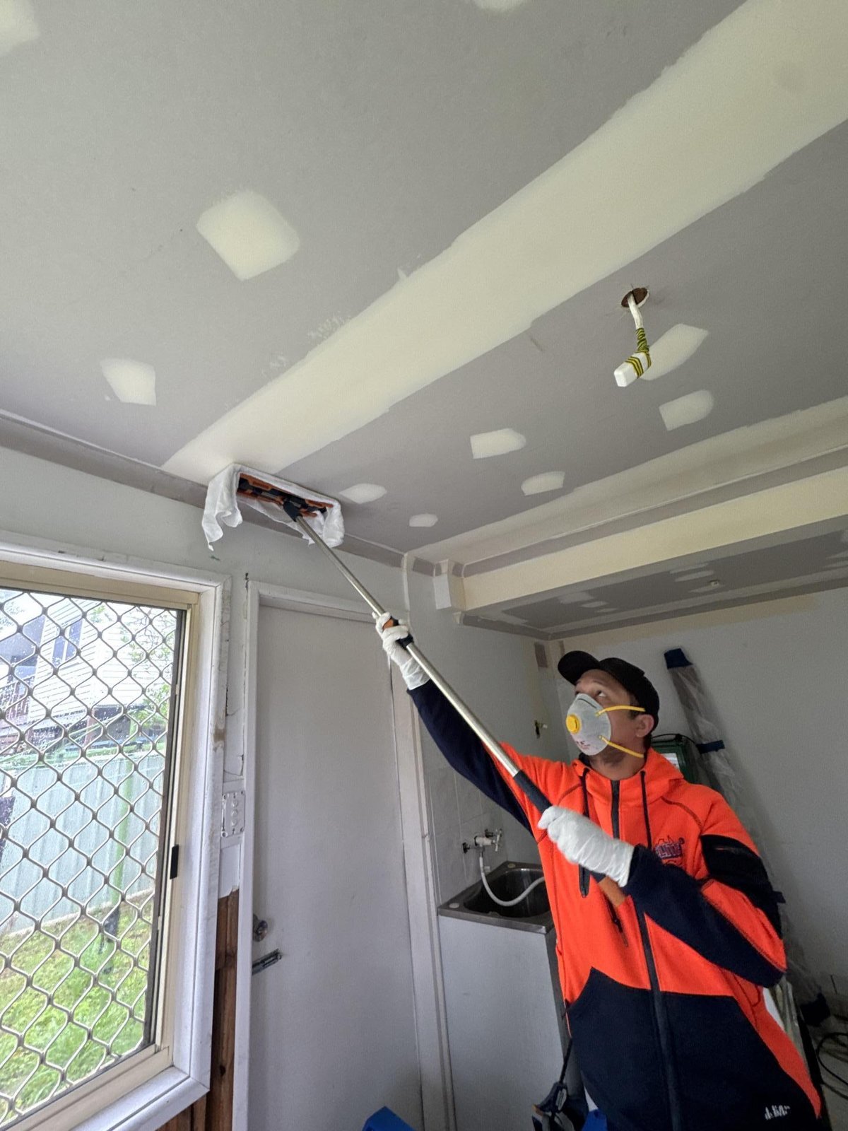 A technician rolls primer onto a ceiling in a small room with a pole roller.