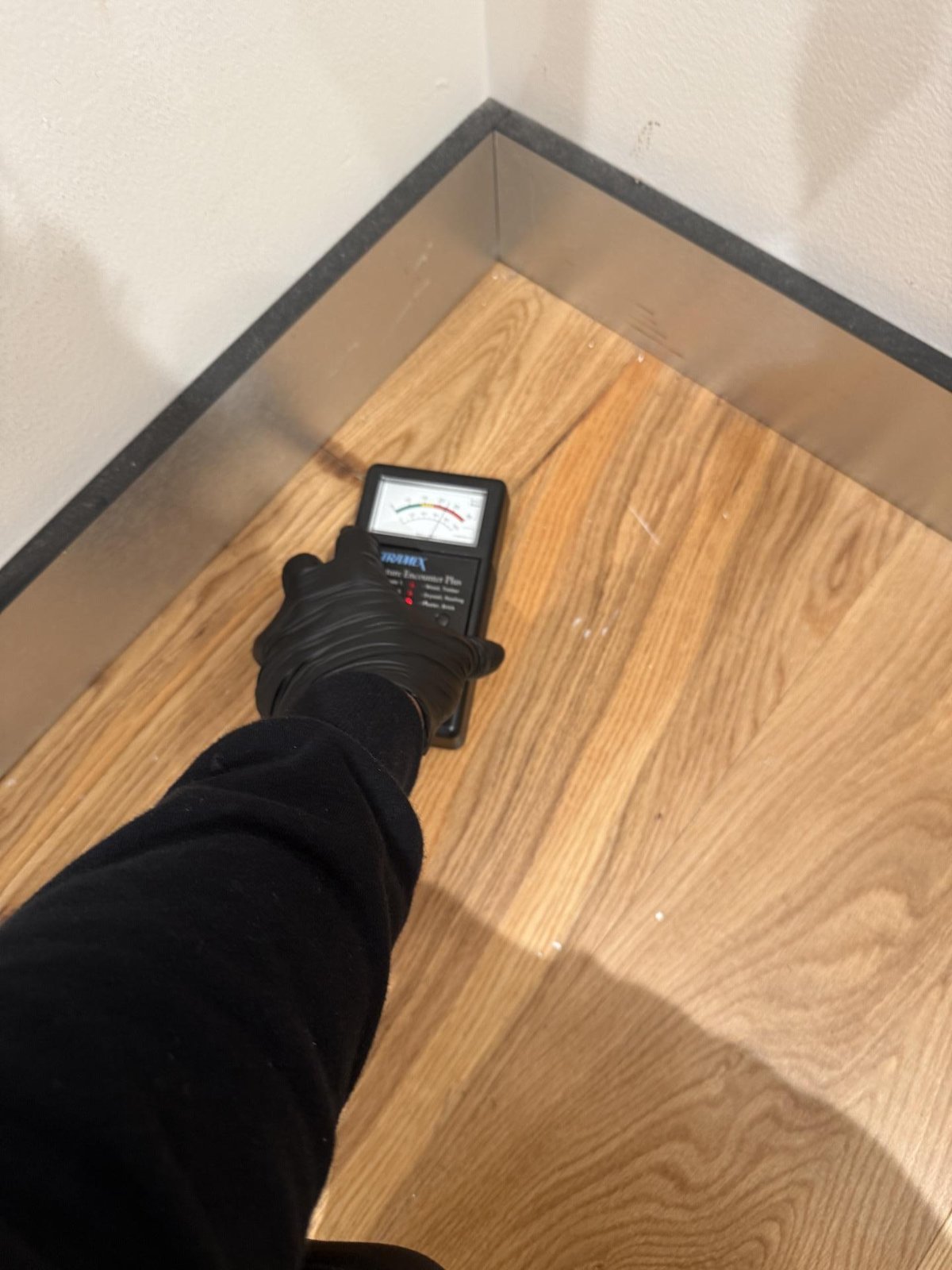 A technician in black gloves tests timber floorboards near a skirting with a moisture meter.