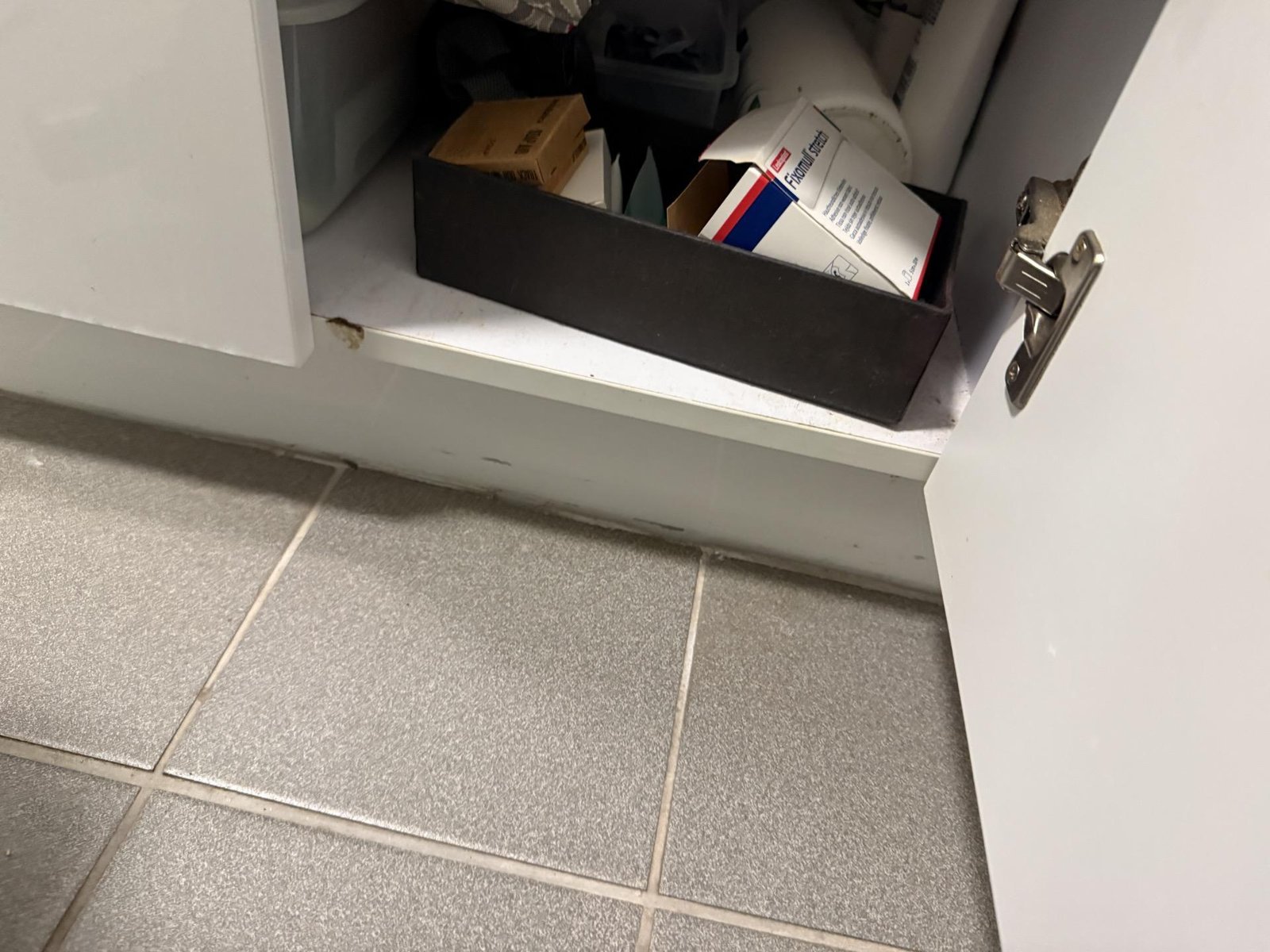 Open vanity cupboard above grey tiled floor, with stored boxes and a small chip on the cabinet base.
