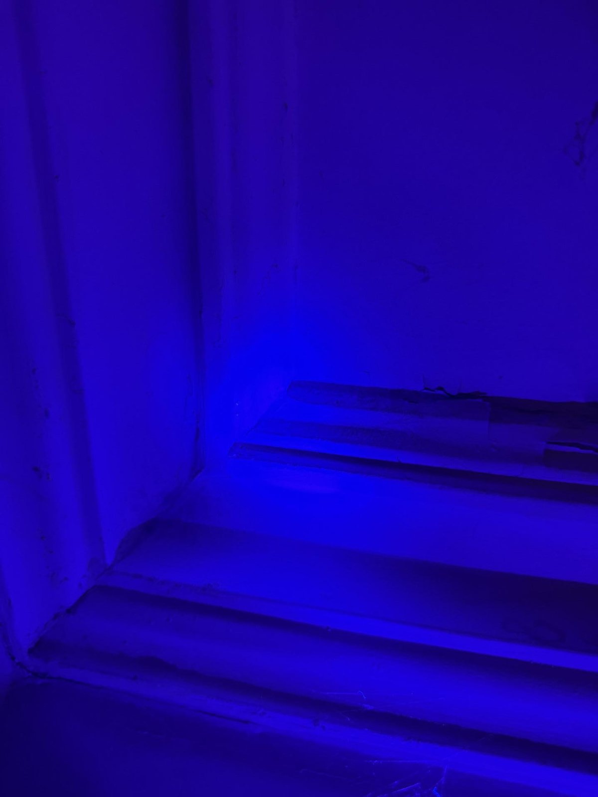 Blue-lit corner of a room with a stepped skirting and damaged wall edges under UV light