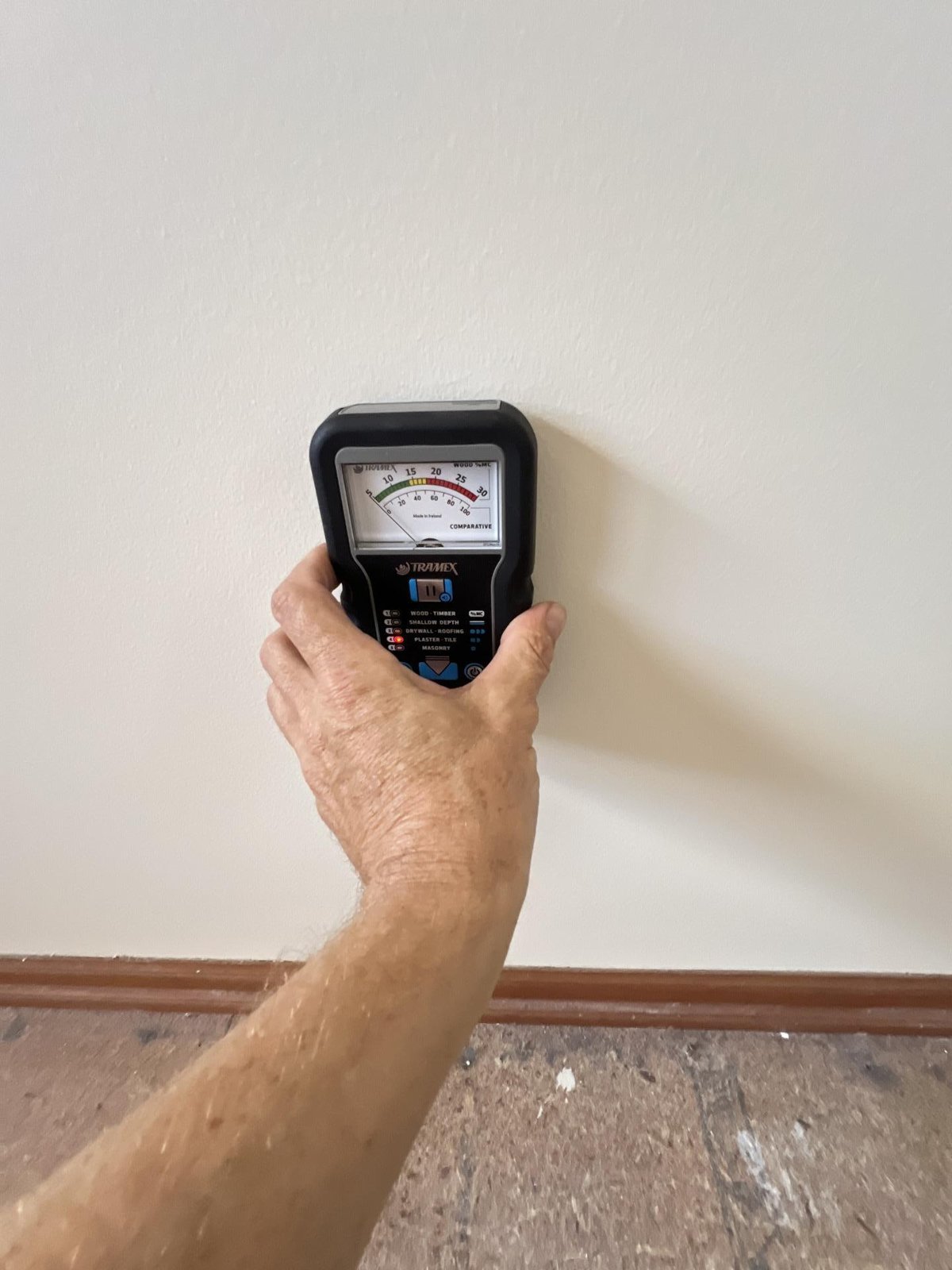 A technician holds a moisture meter against a wall above a stripped skirting board.
