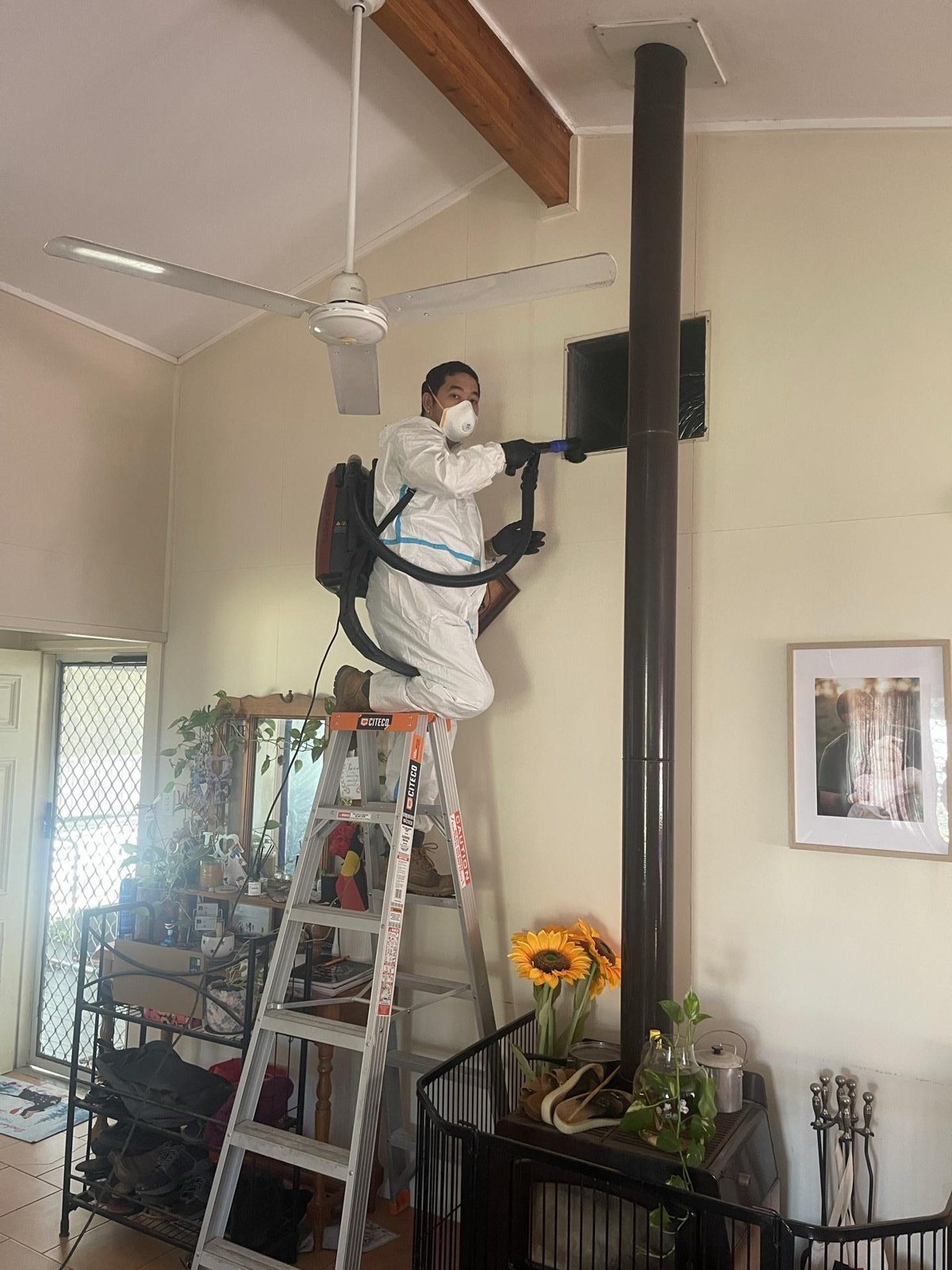 A technician in a white coverall and respirator on a ladder, using a hose to treat a wall vent.