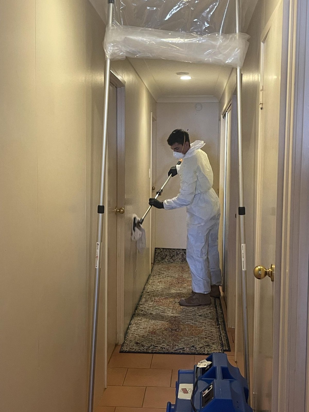 A technician in white protective suit cleaning a hallway wall, with plastic sheeting overhead and a blue air mover at the floor.