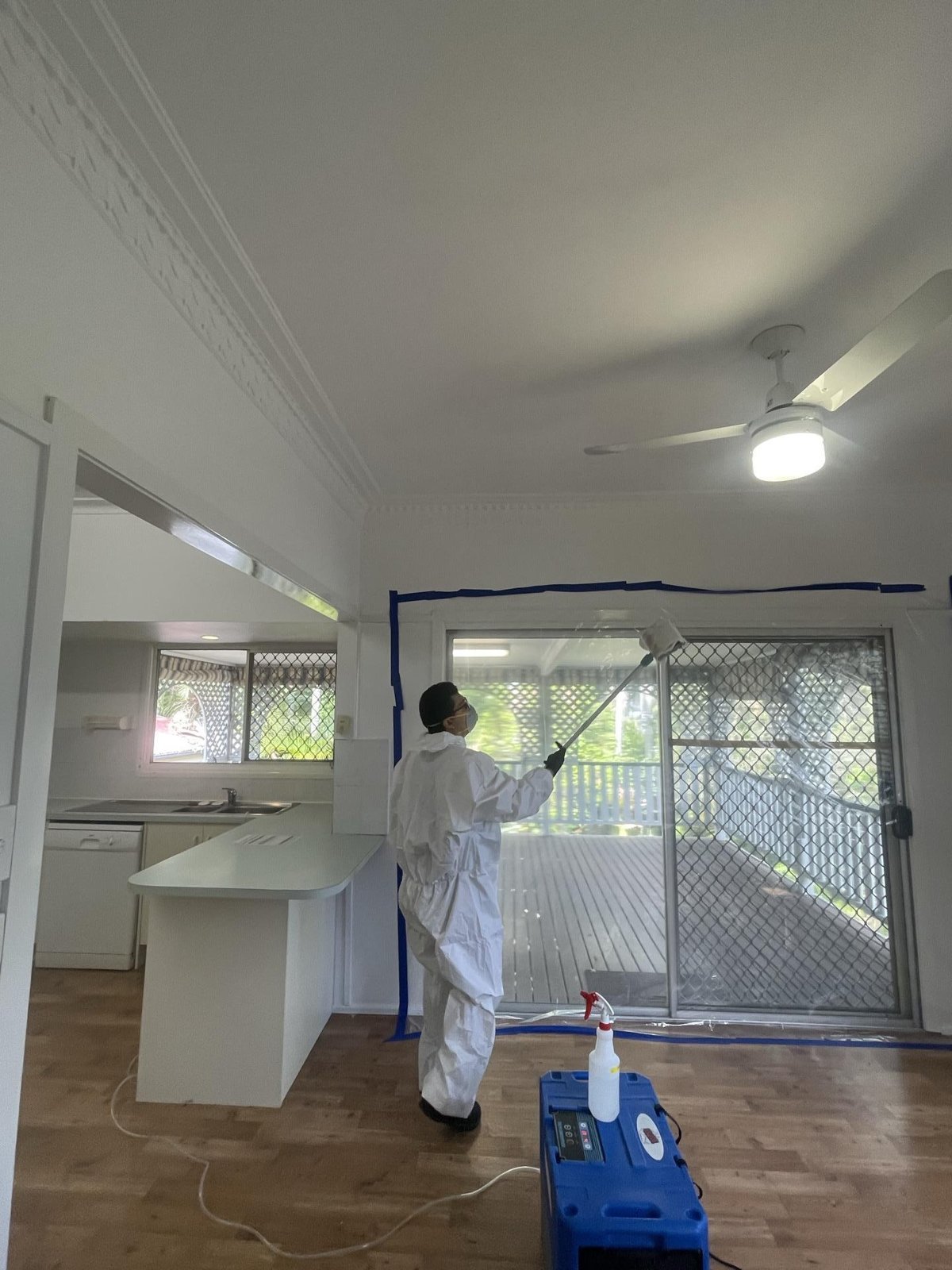 A technician in white protective overalls cleans a ceiling with a long-handled tool, with a blue dehumidifier on the floor.