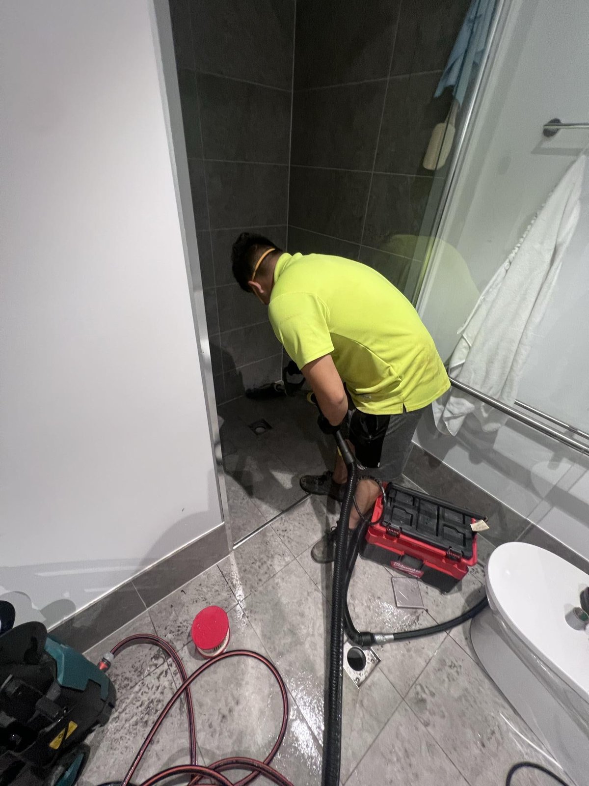 A technician uses a wet vacuum hose on a wet tiled shower floor, with a red toolbox nearby.